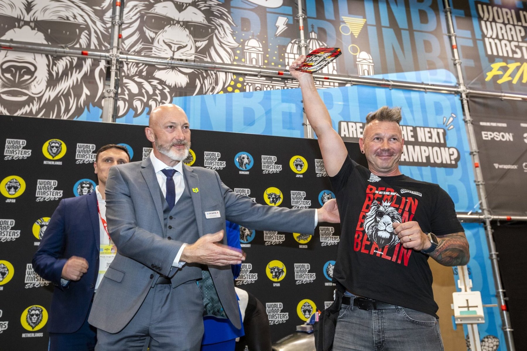A smiling Mike Szwacki raises his prize at the FESPA Wrap Masters Finals, congratulated by an official in a suit.