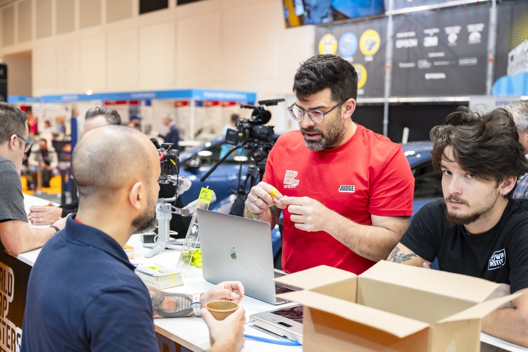 A judge at the World Wrap Masters competition examines material over a laptop, with competitors nearby.