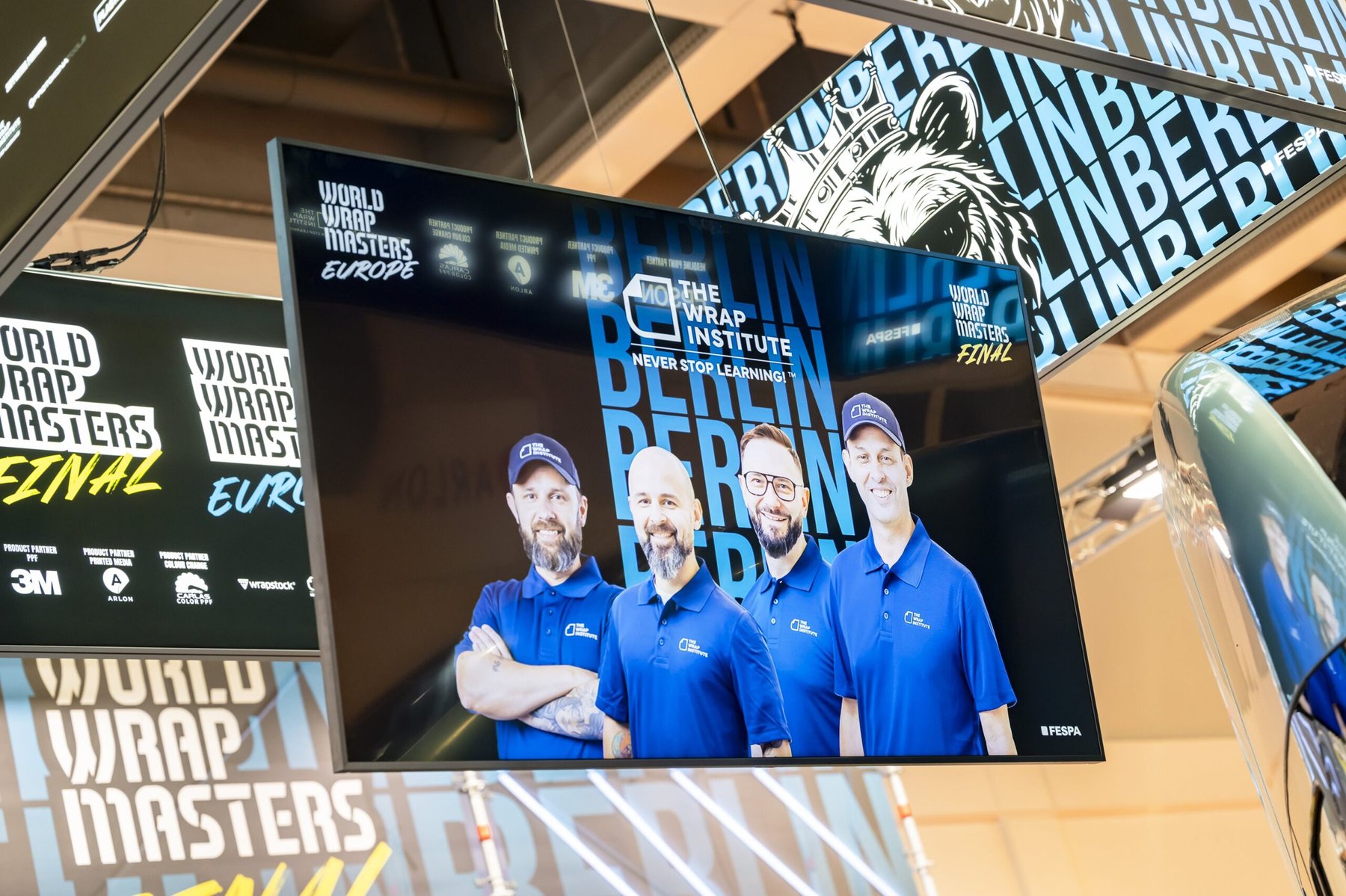 Digital screens at FESPA Wrap Masters, featuring four smiling men from The Wrap Institute.