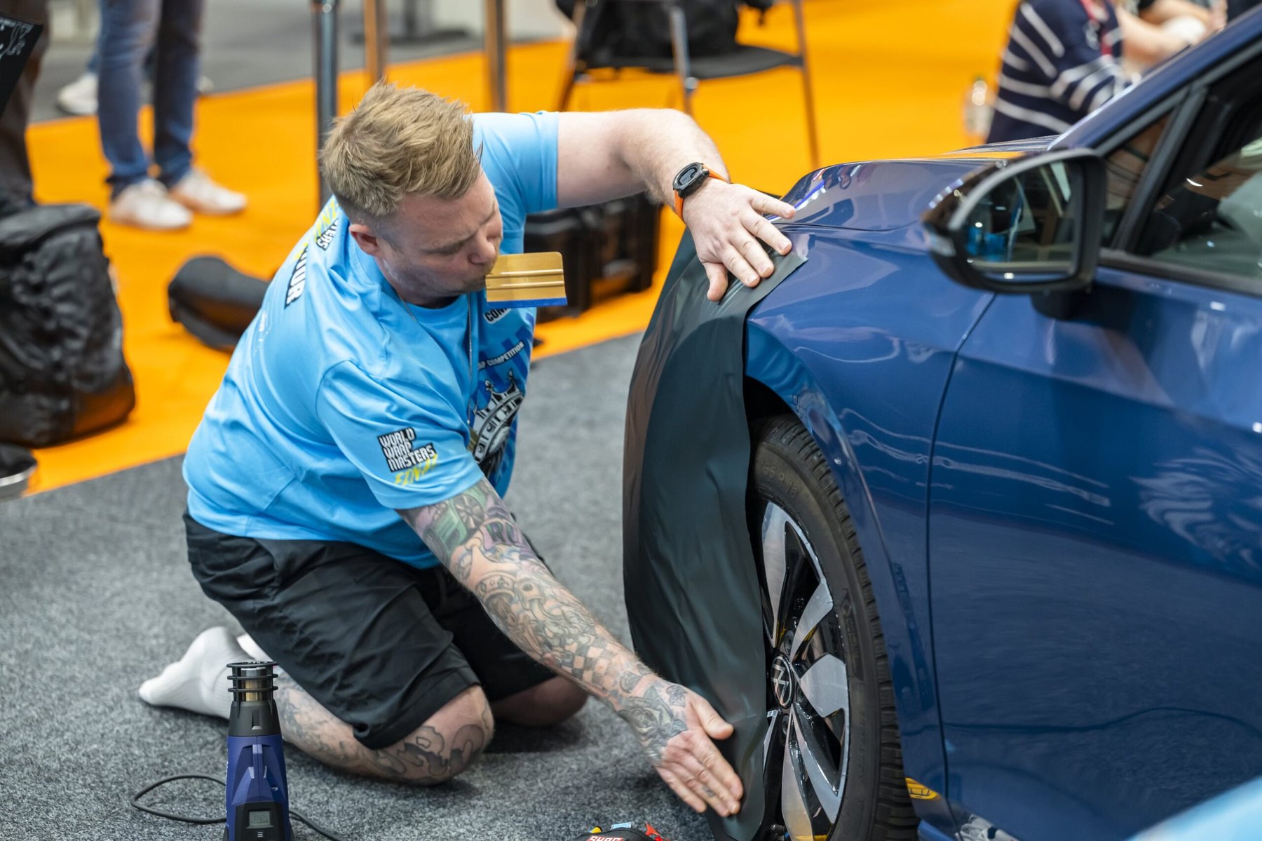 A competitor applies a dark vinyl wrap to the front wing of a blue car during the Wrap Masters competition.