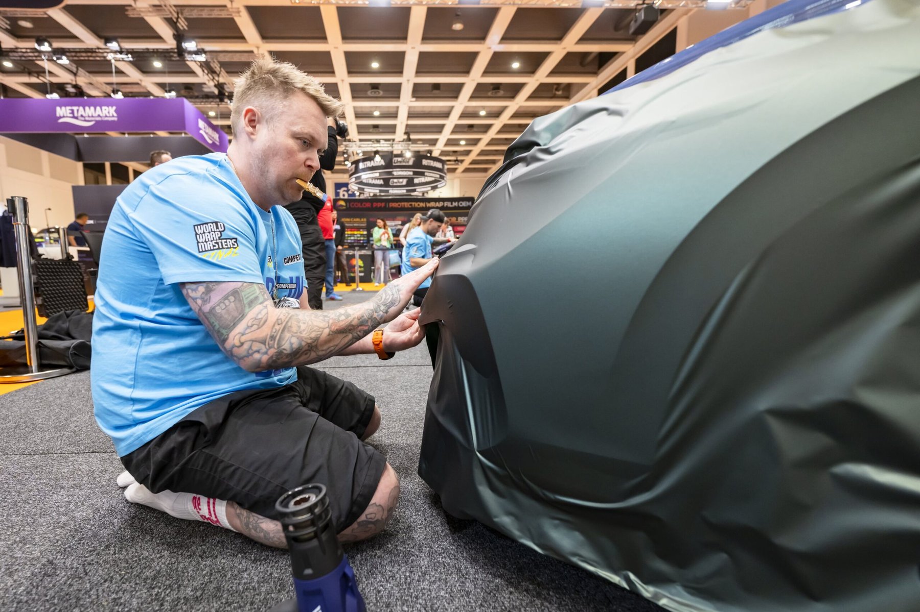 Tattooed man, wearing a World Wrap Masters shirt, kneels while applying dark vinyl film to a vehicle.