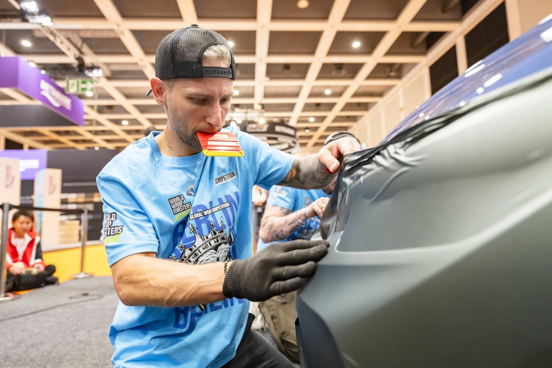 A FESPA Wrap Masters competitor applies grey vinyl film to a car, holding a squeegee in his mouth.