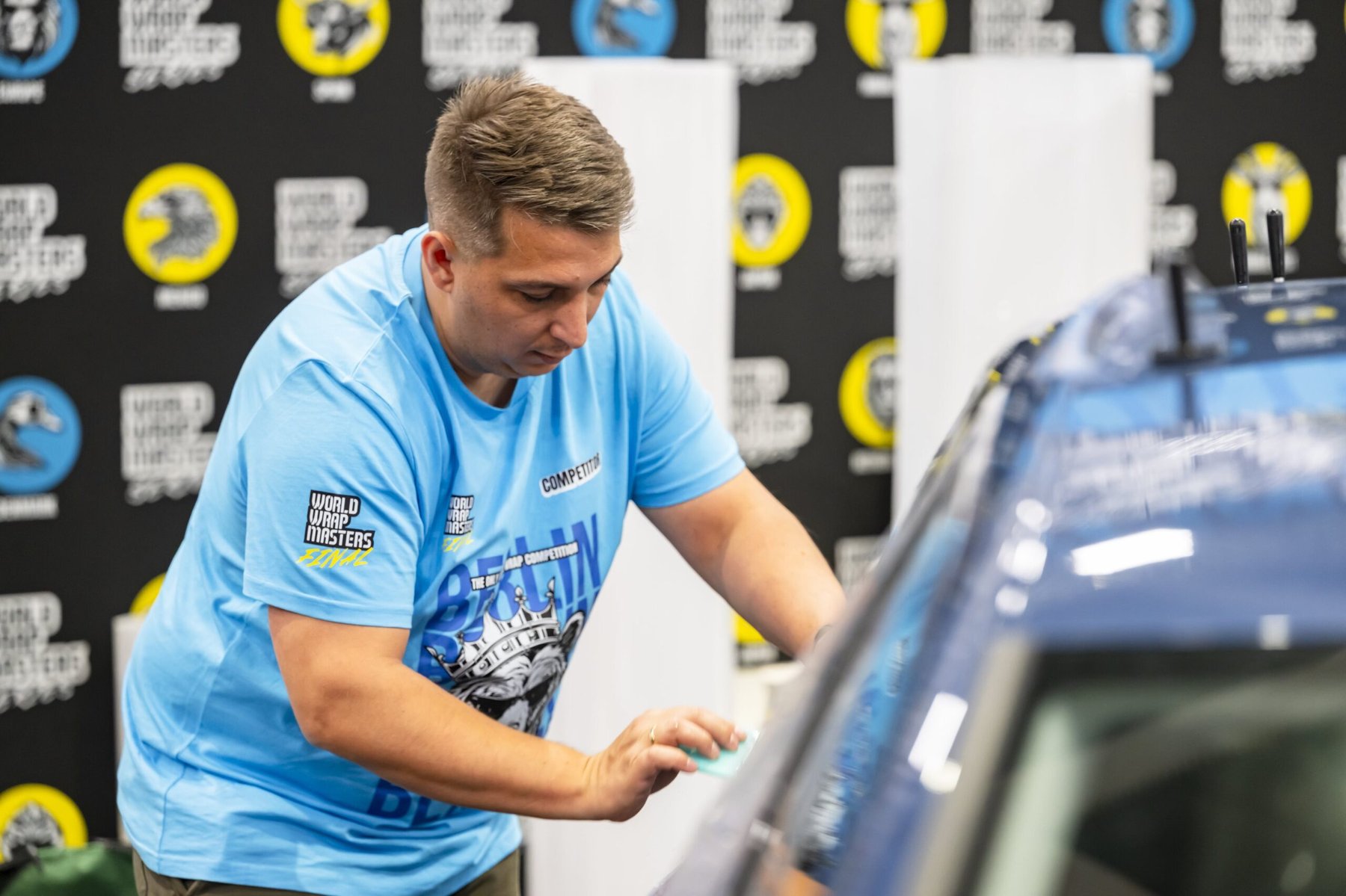 A competitor applies vinyl wrap to a dark vehicle using a squeegee at the World Wrap Masters competition.