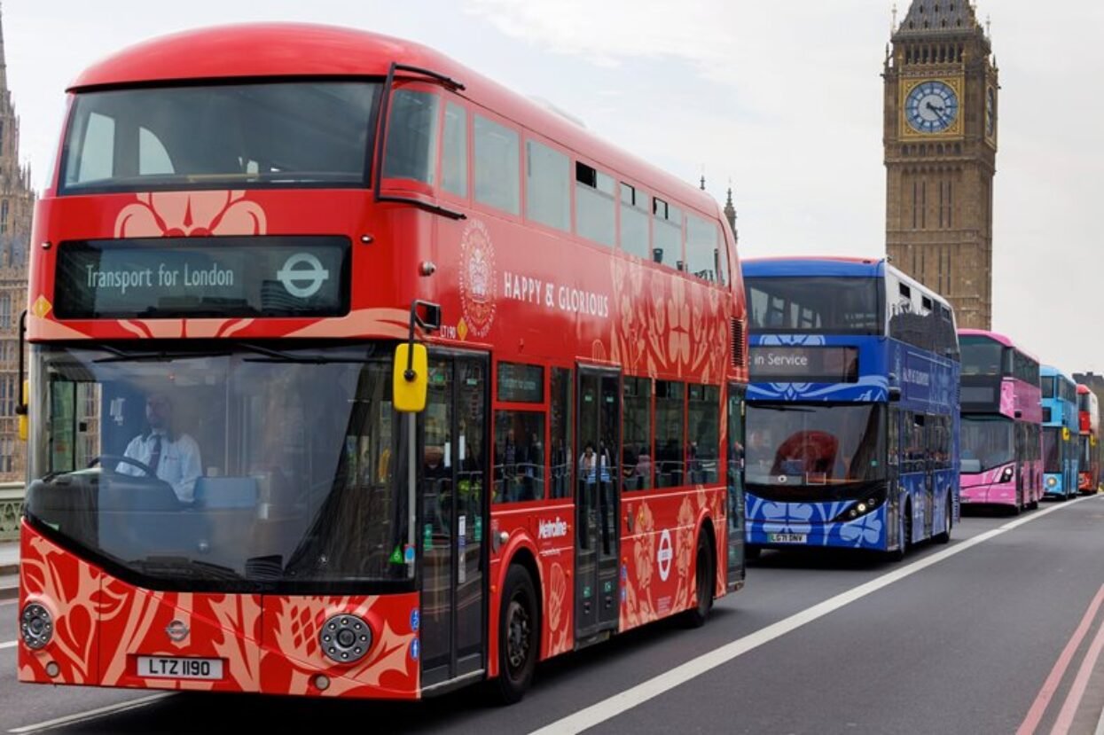Custom wrapped double-decker buses, red, blue, and pink, queuing near Big Ben, London.
