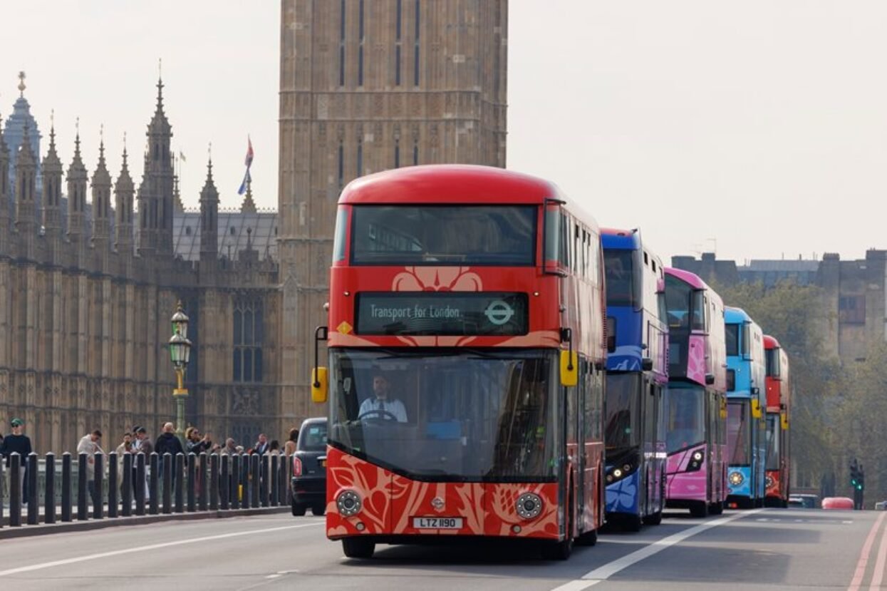 Red, blue, and pink wrapped double-decker buses driving past the Palace of Westminster, London.