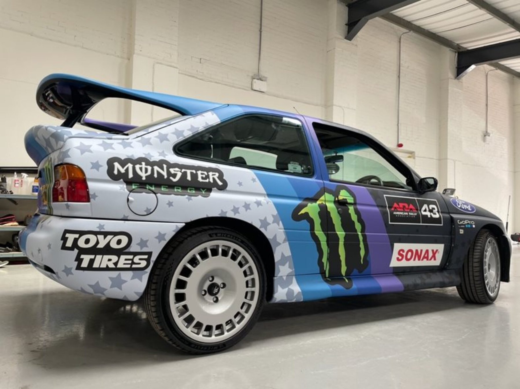 Competition rally car, wrapped in blue, purple, and grey vinyl, featuring Monster Energy and Toyo Tires branding.