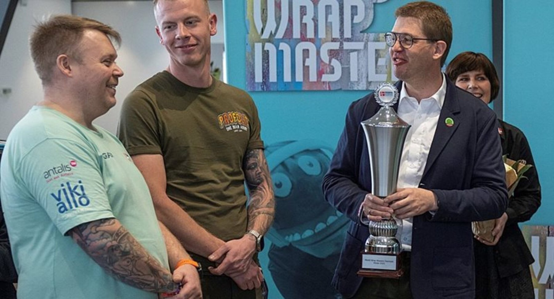 A man in a suit holds the Wrap Masters trophy, congratulated by two competitors.