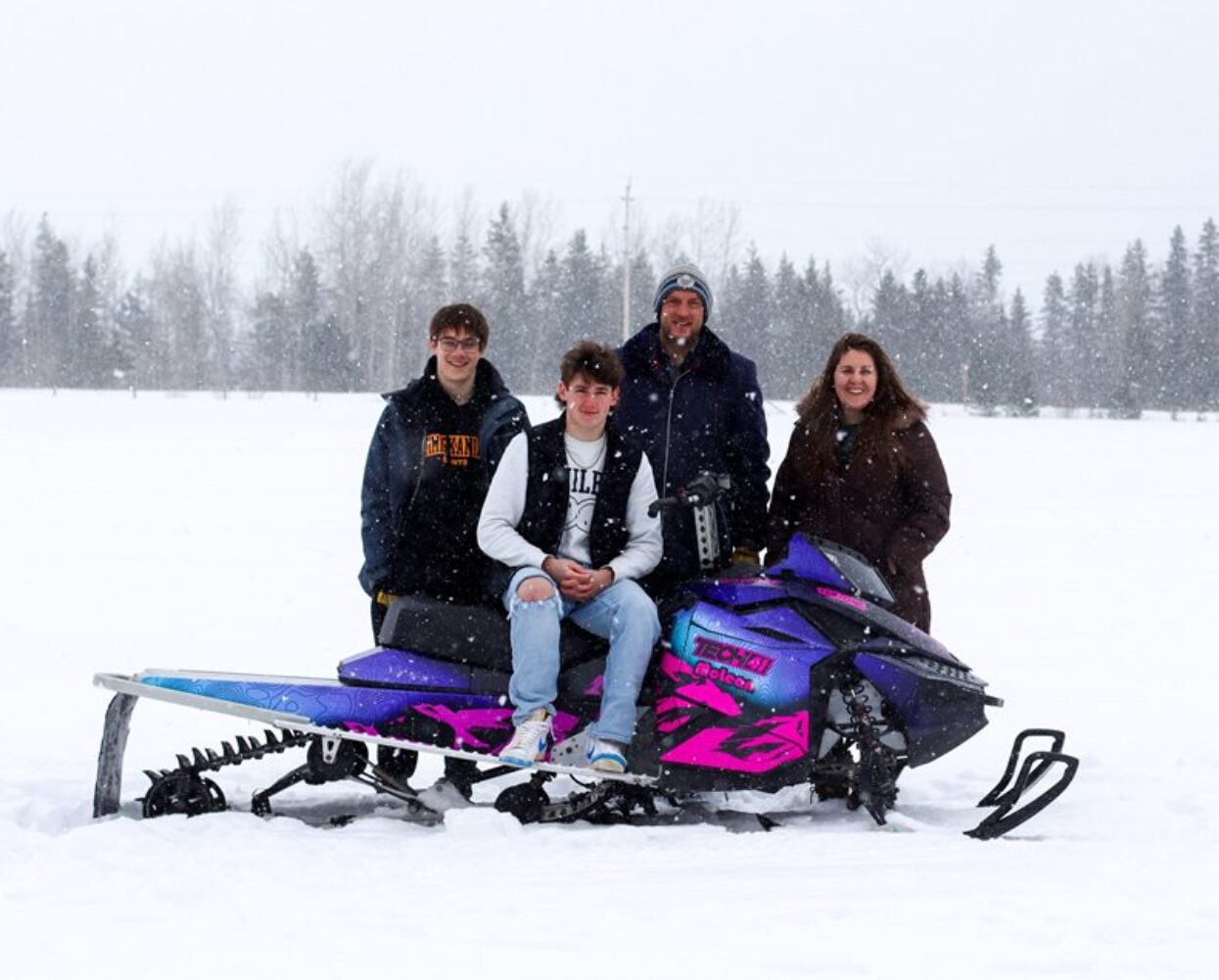 Four people stand with a custom wrapped purple and pink snowmobile in a snowy field.