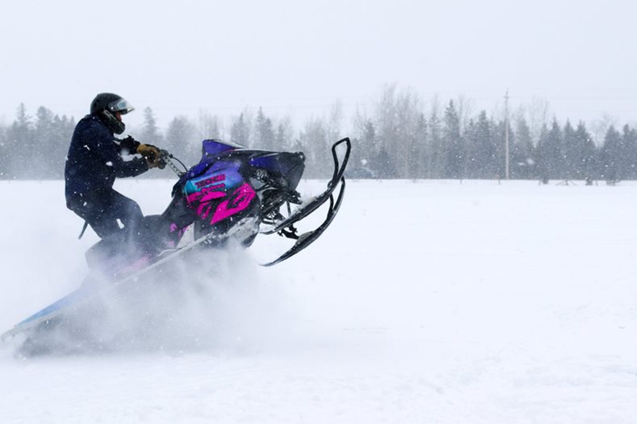 A rider on a purple and pink wrapped snowmobile kicking up snow in a winter field.