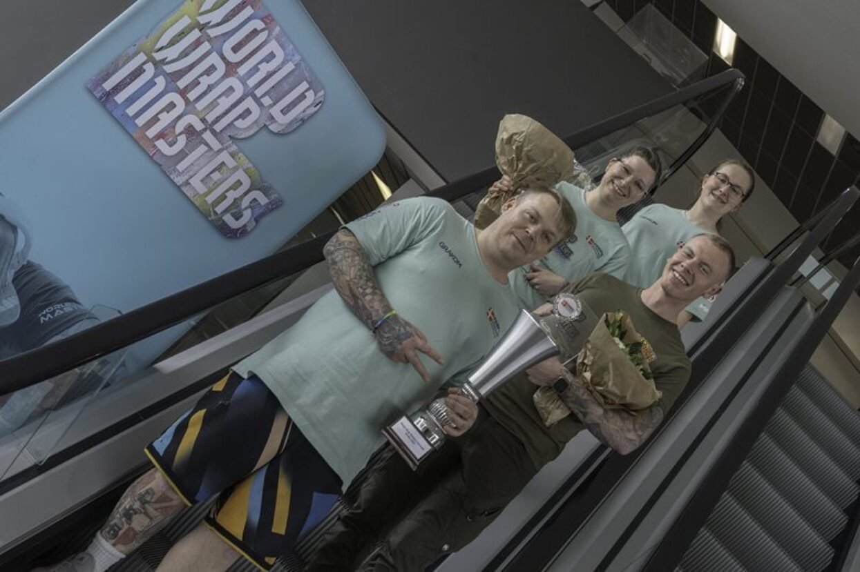Four smiling winners holding a trophy and flowers on an escalator at the World Wrap Masters competition.