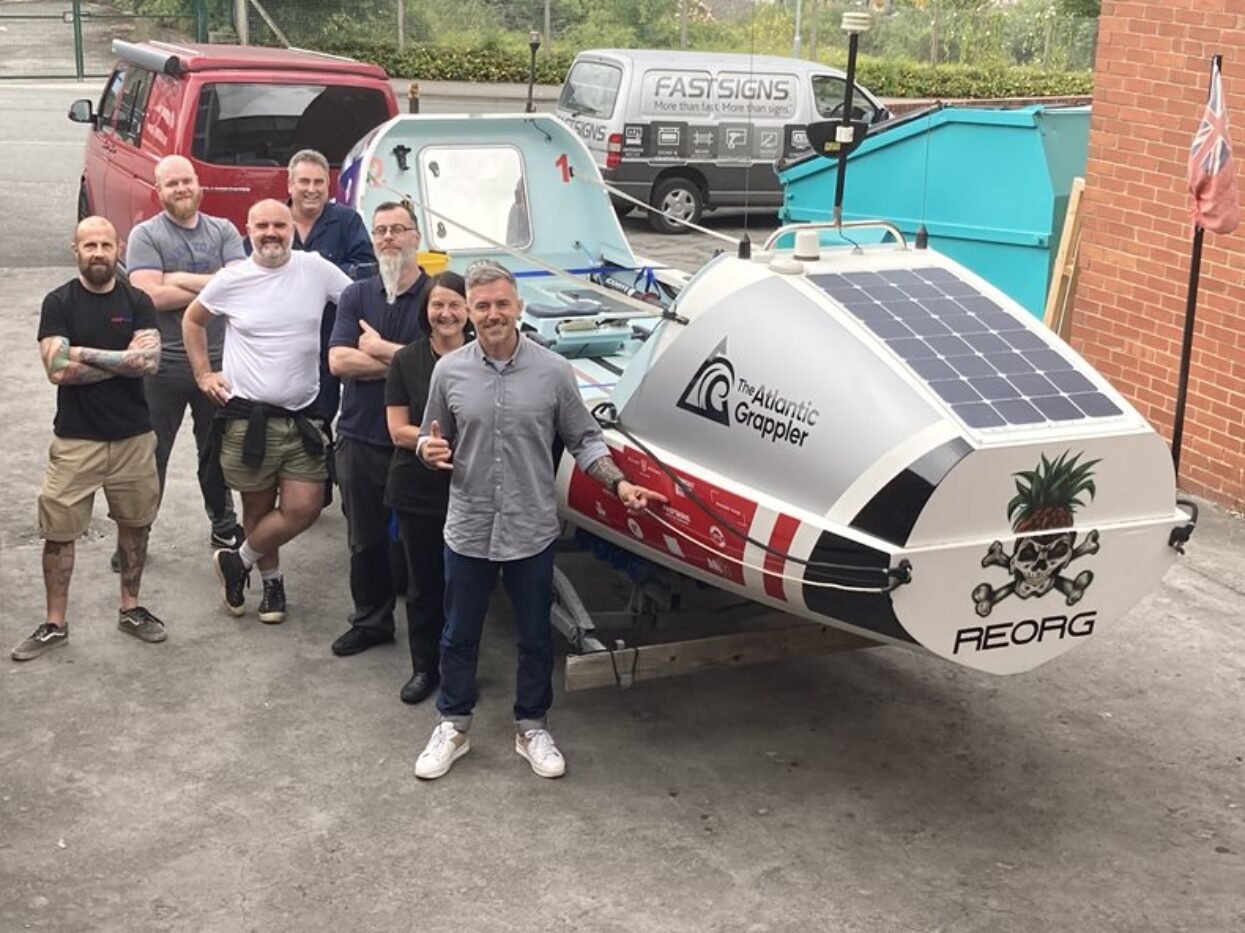 Seven people standing next to The Atlantic Grappler, a small, wrapped boat with solar panels.