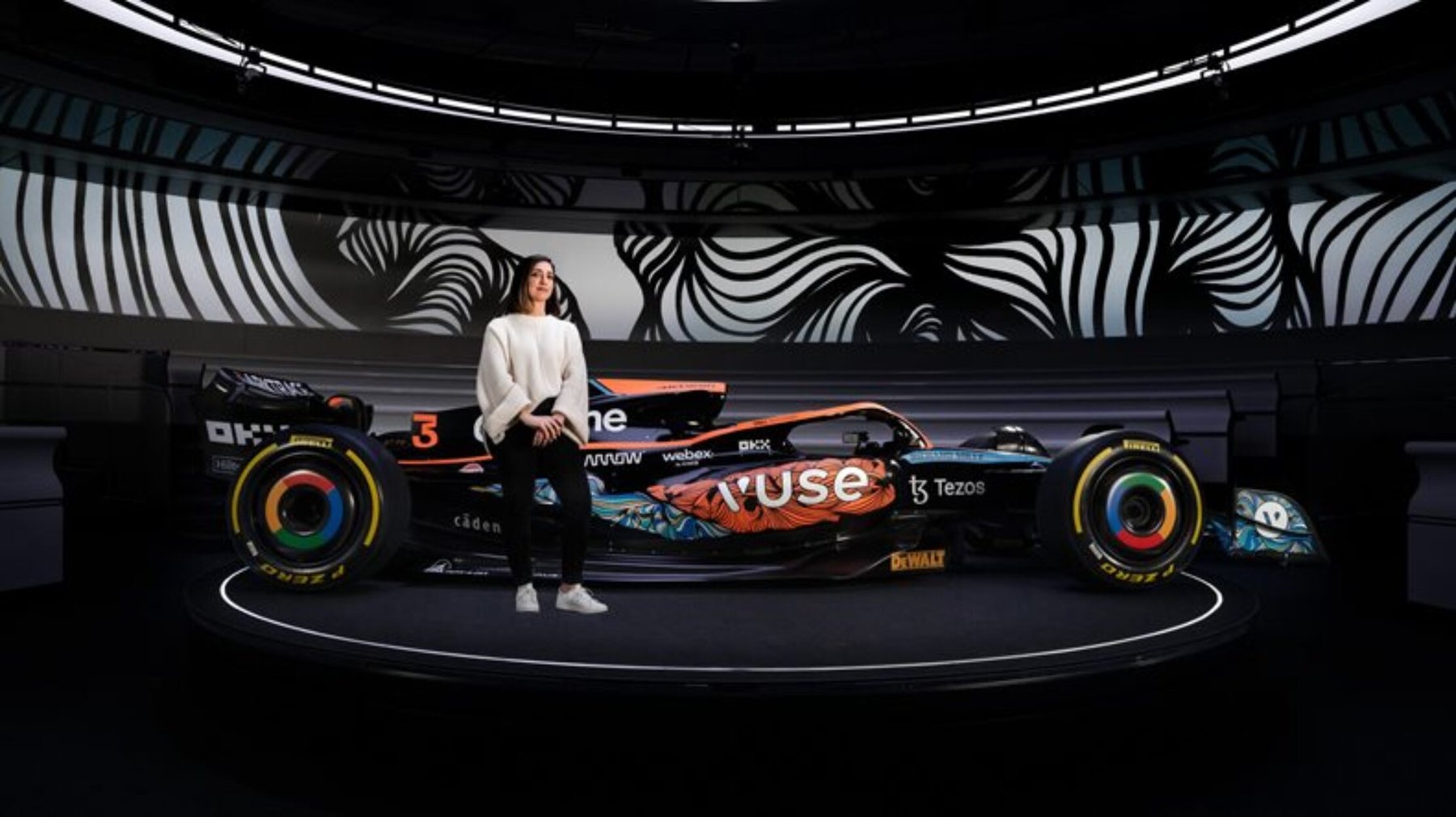 Woman stands next to a custom-wrapped black and orange racing car displayed on a dark stage.