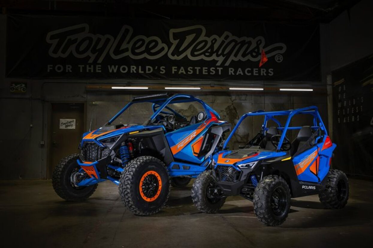 Two blue and orange wrapped Polaris RZR vehicles in a garage, promoting Troy Lee Designs.