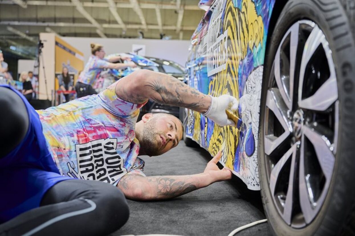 A vehicle wrapper, lying down, carefully applies a colourful tiger-stripe vinyl to the lower car panel.