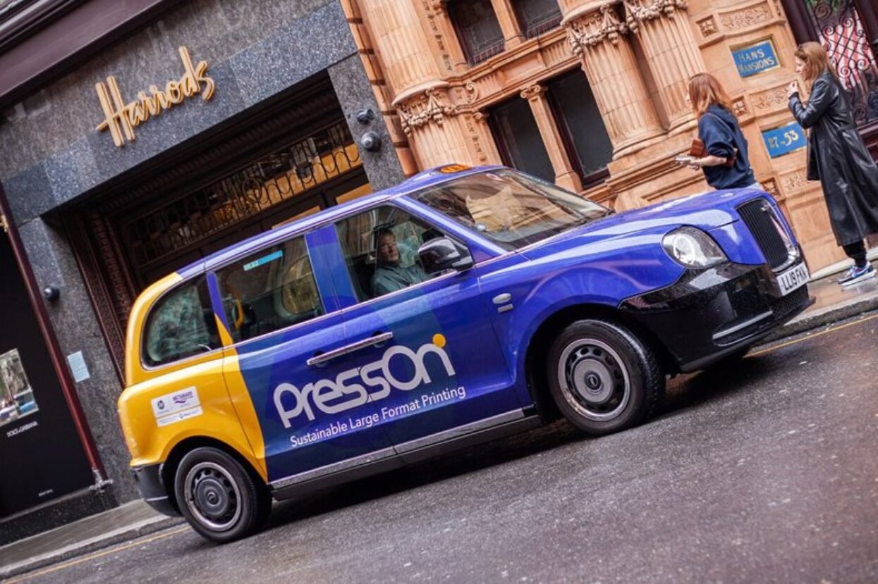 London black cab wrapped in blue and yellow PressOn advertising, parked outside Harrods.
