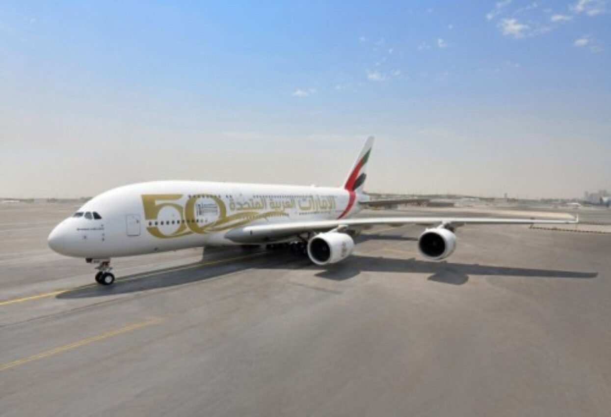 Emirates A380 aircraft with a gold 50th anniversary livery wrap, parked on an airport runway.
